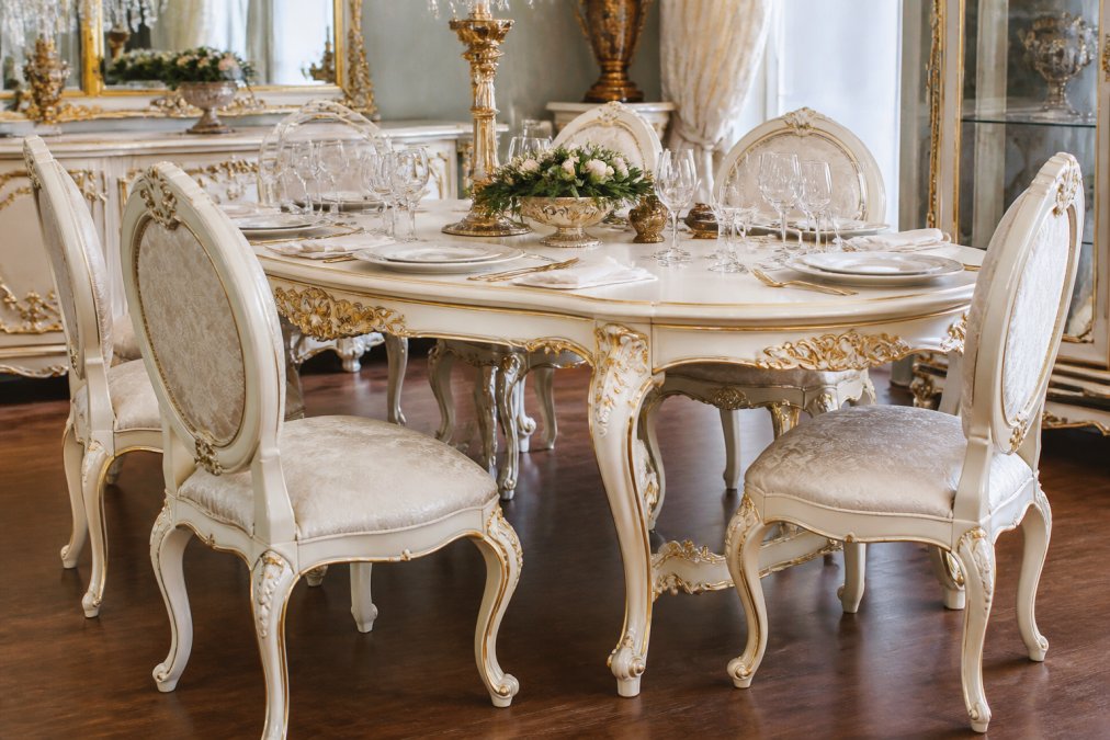 Handgefertigter Rokoko-Esstisch mit geschwungenen Cabriole-Beinen und feinen Rocaille-Ornamenten in Weiß und Gold, eingedeckt für ein elegantes Dinner in einem hellen, luxuriösen Esszimmer mit Kronleuchter und klassischen Elementen