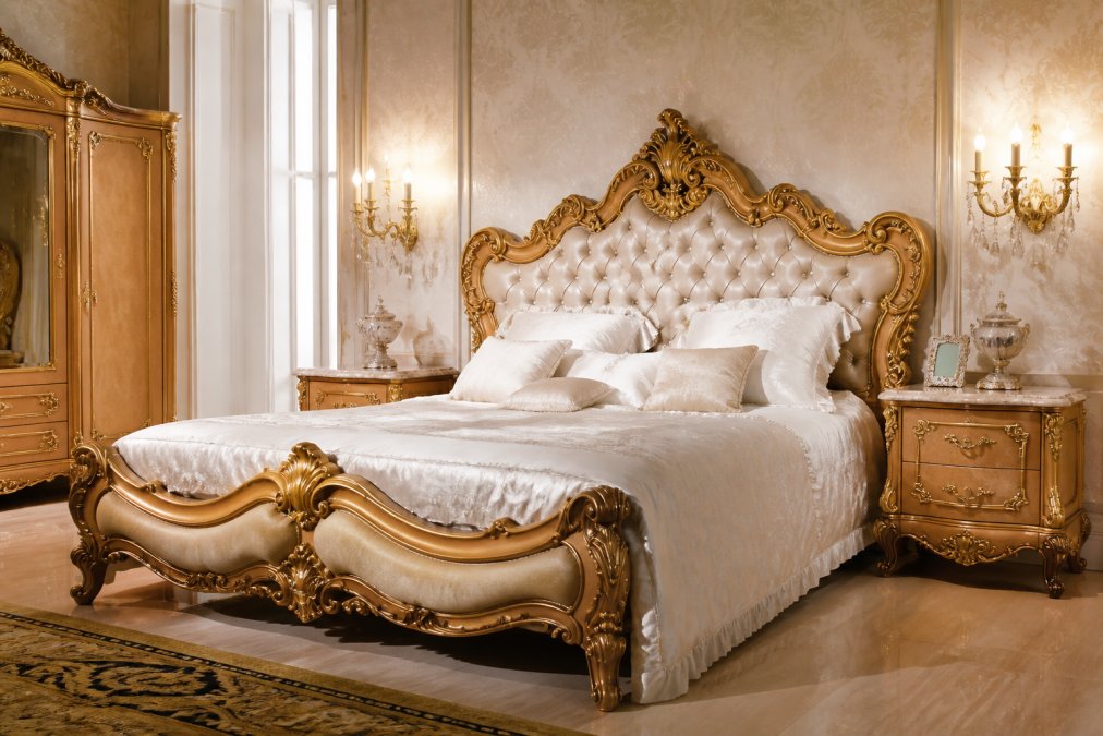 Elegantes Schlafzimmer mit prunkvollem Barockbett als zentrales Möbelstück, Massivholz-Betthaupt mit handgeschnitzten Verzierungen in Goldapplikationen, umgeben von klassischen Nachttischen mit Marmor-Oberflächen, goldenen Wandleuchten mit Kristallbehang, edlen Textilien in Creme und Weiß, warme indirekte Beleuchtung, helle Wände mit feinen Mustertapeten