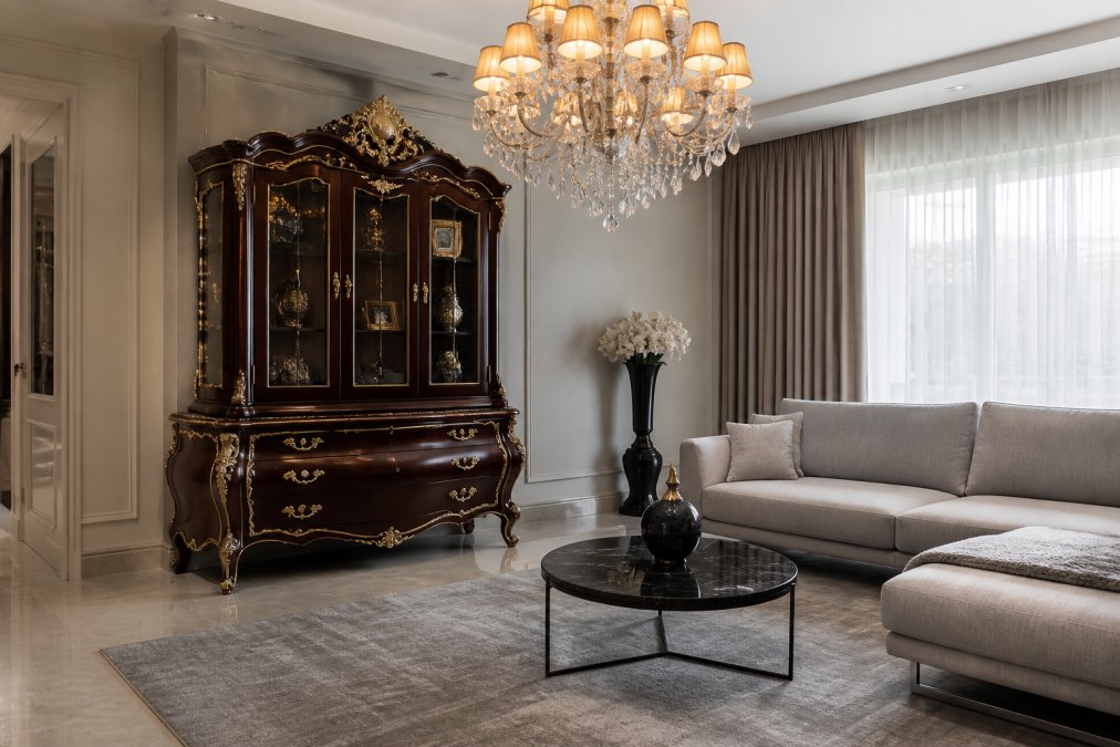 Stilvolles Wohnzimmer mit dunklem Mahagoni-Barockschrank mit bronze-goldenen Verzierungen und geschwungenen Beinen, modernes hellgraues Sofa daneben, eleganter Kronleuchter mit warmer Beleuchtung von oben, minimalistischer grauer Teppich