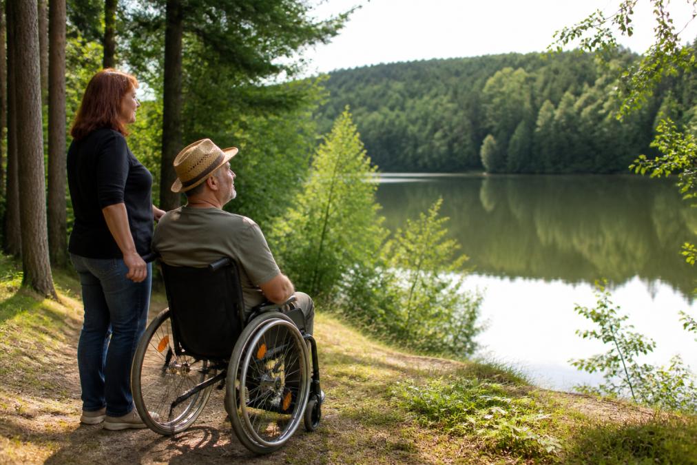 Osoba na invalidním vozíku si u klidného jezera ve Waldviertelu vychutnává výhled, vedle ní stojí doprovod, zelená lesní krajina, měkké odpolední světlo