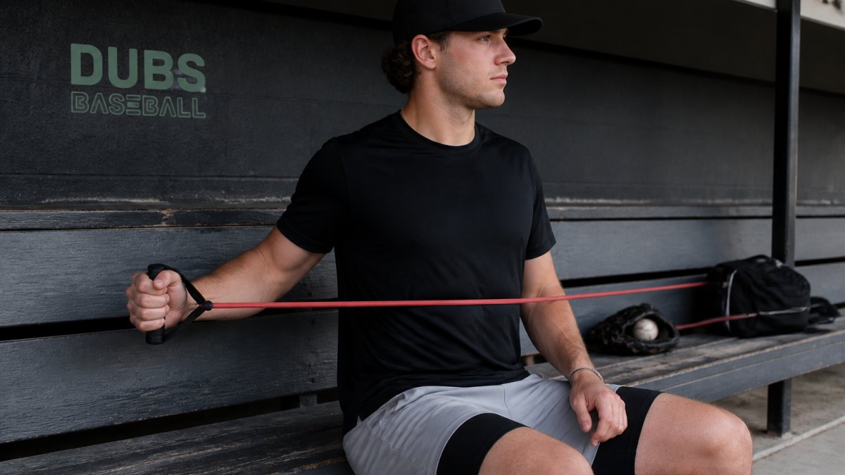 Baseball pitcher seated on dugout bench performing light banded external rotation at side after a throwing session, showing relaxed posture and controlled tempo