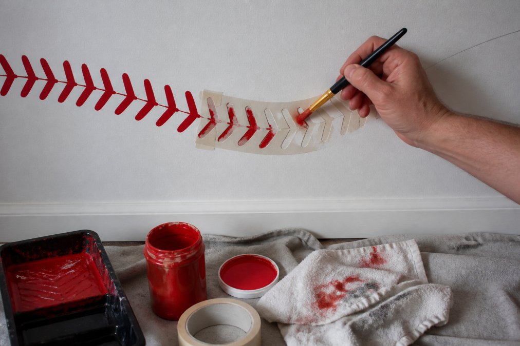 Close-up of a hand holding a paintbrush carefully applying red baseball stitches along a stencil line on a white wall, with paint supplies and drop cloth visible on the floor