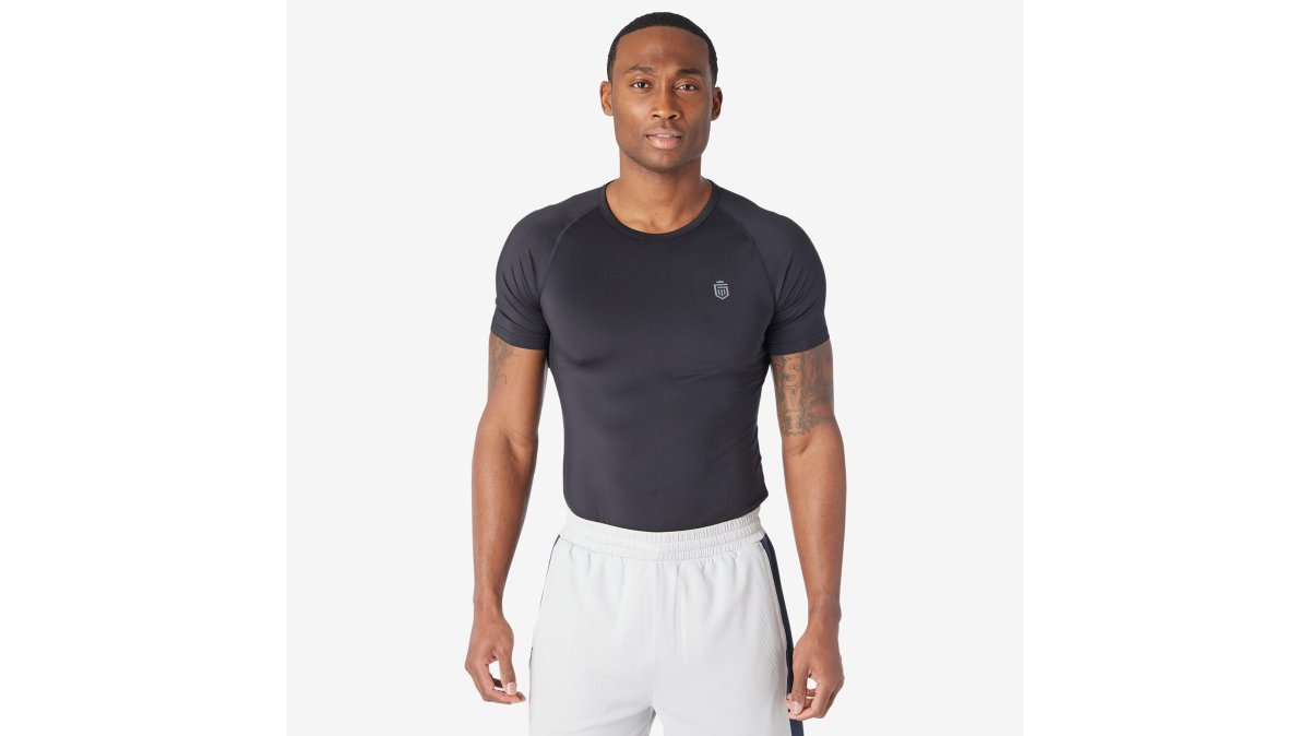 An athlete mid-workout in a gym wearing a lightweight gray Baselayer Tee, sweat visible on the mesh ventilation panels along the chest and back, dramatic side lighting highlighting the fabric's moisture-wicking technology