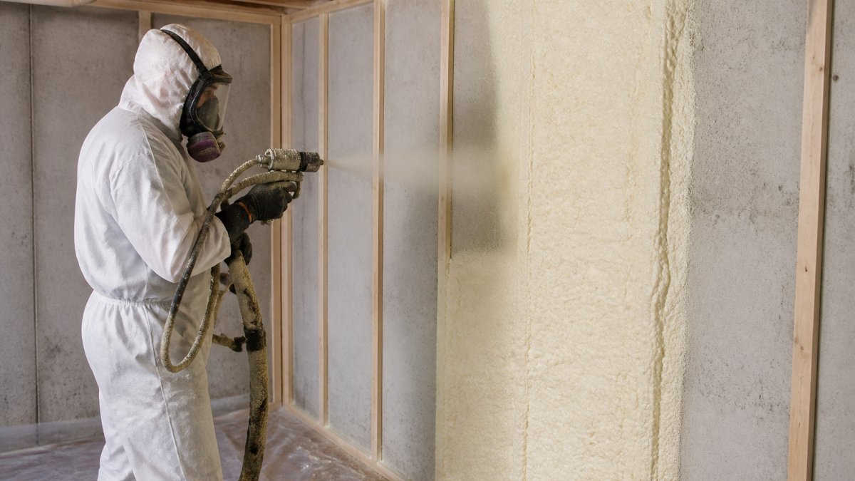 Professional contractor in protective gear and respirator applying closed-cell spray foam insulation to a basement foundation wall, with foam expanding and curing visible