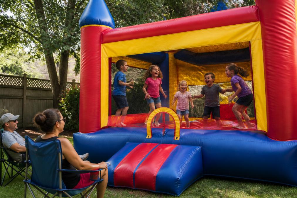 Group of children laughing and playing inside a colorful bounce house at a backyard party, with adults supervising from nearby lawn chairs in afternoon sunlight, trees and residential fence visible in background