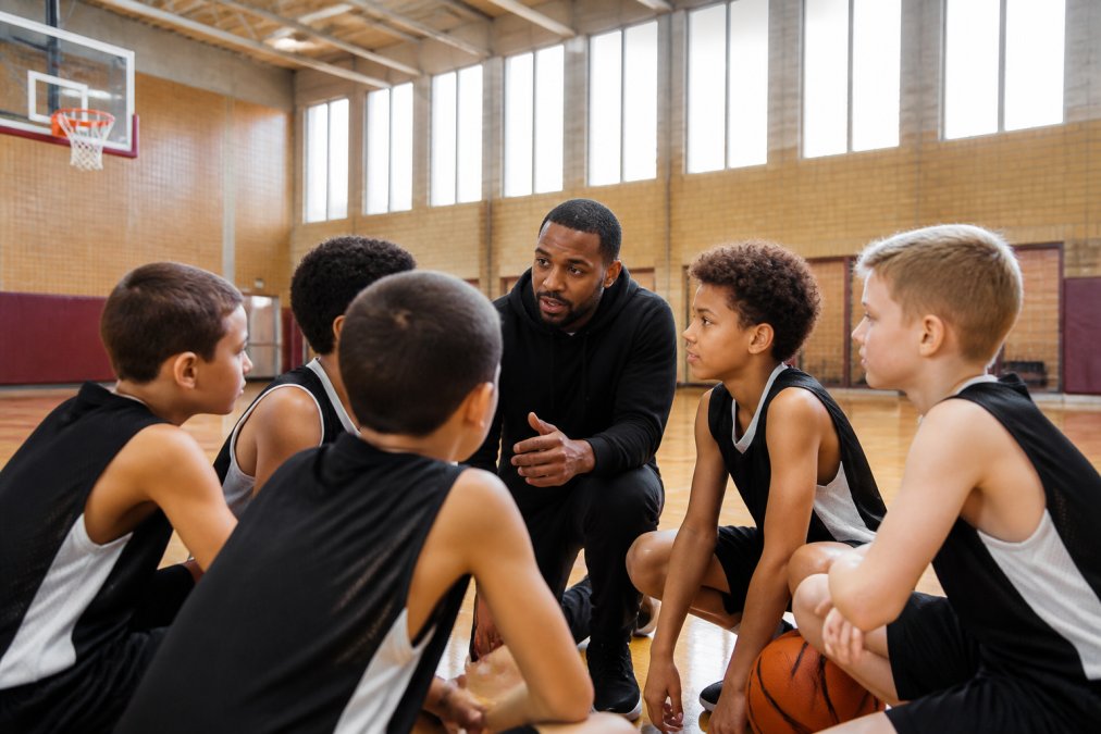 Young basketball players (ages 8-10) gathered in a huddle with their coach, demonstrating teamwork and communication during a practice session in a gymnasium with natural light streaming through tall windows