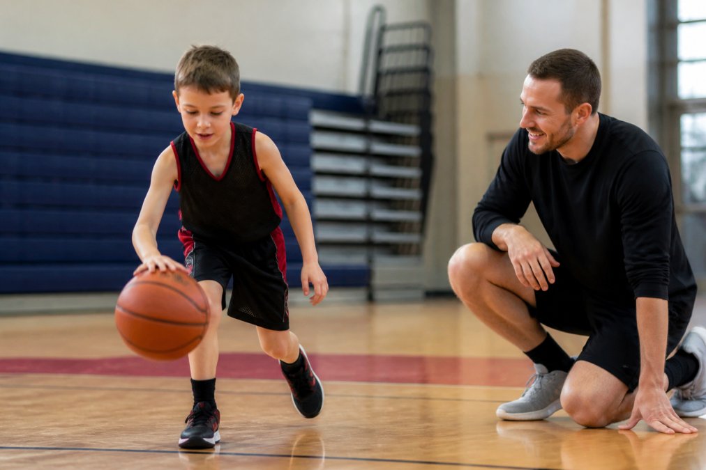 A young child around 6 years old, wearing a basketball jersey, dribbling a ball on an indoor community court with a supportive coach kneeling nearby and smiling
