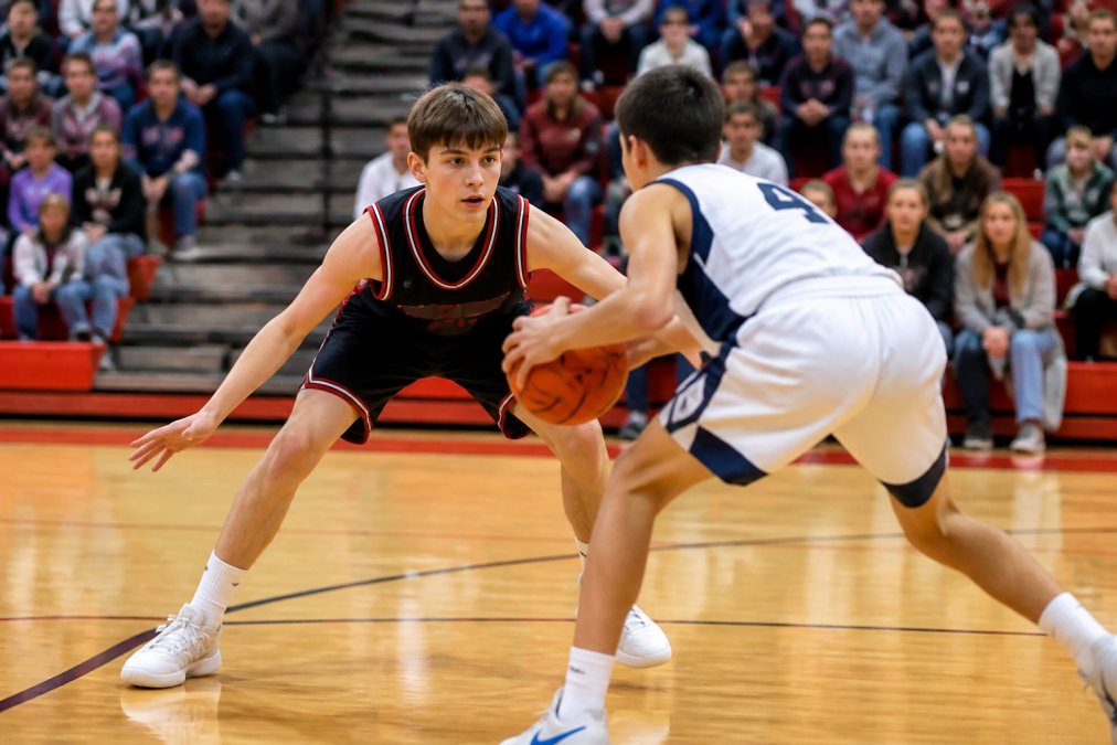 Young basketball player in defensive stance during competitive game, focused expression, opponent attempting a drive, bright gymnasium setting with spectators visible