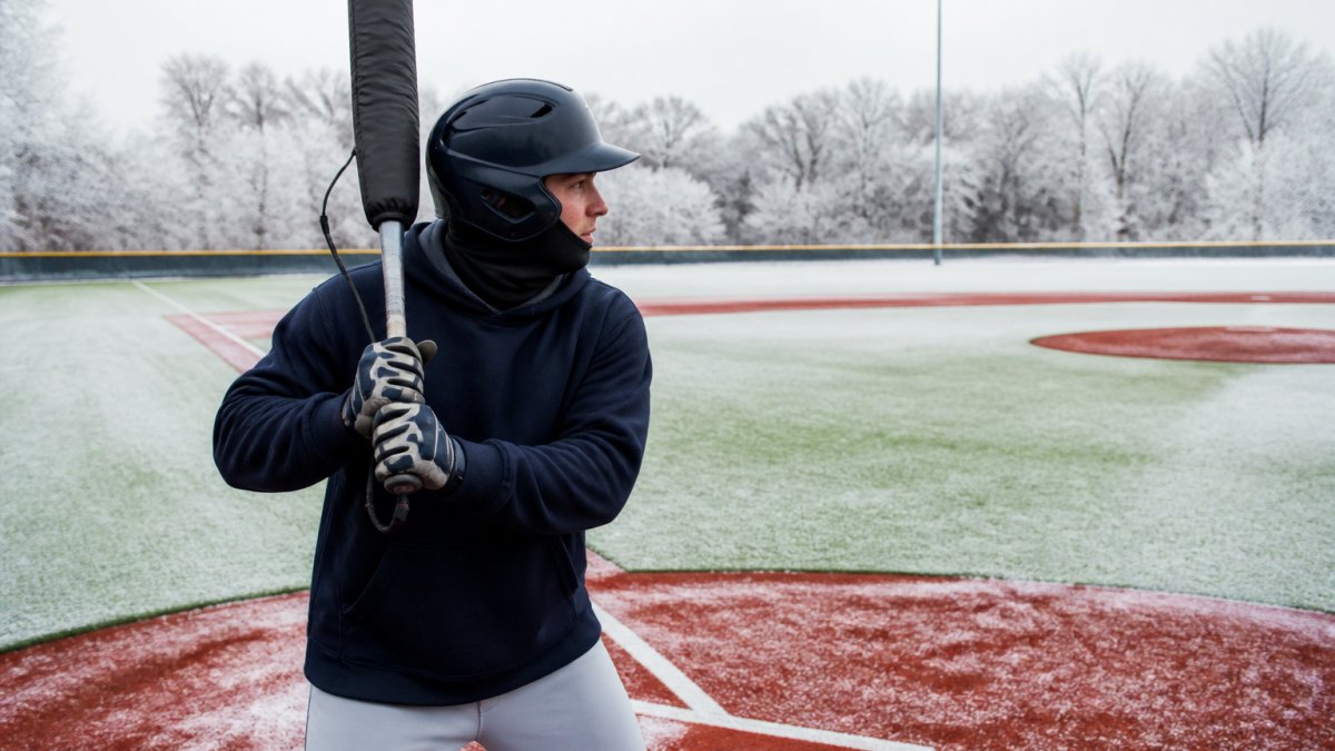Baseball player in cold weather gear using a heated bat sleeve on a frosty field