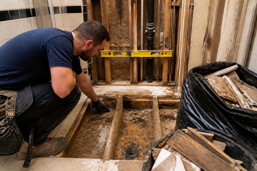 Renovator inspecting a bathroom subfloor for water damage during a shower remodeling project.