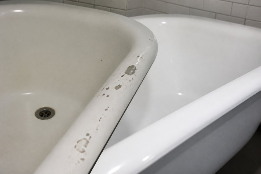 Close-up of a bath surface showing visible chips, scratches, and discolouration on aged white enamel, contrasted with a section of pristine new porcelain fixture in bright bathroom lighting