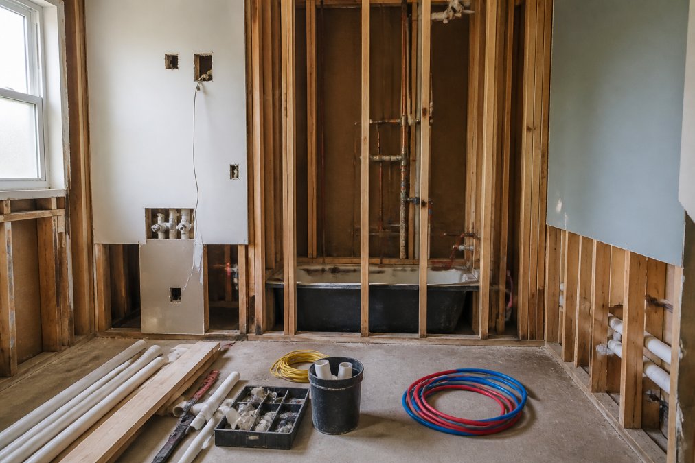 A bathroom mid-renovation showing exposed rough-in plumbing and electrical wiring, with drywall partially removed and construction materials organized on the floor
