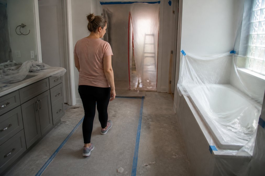 A homeowner walking through a bathroom with plastic dust barriers and protective coverings on fixtures, showing the reality of daily living during renovation
