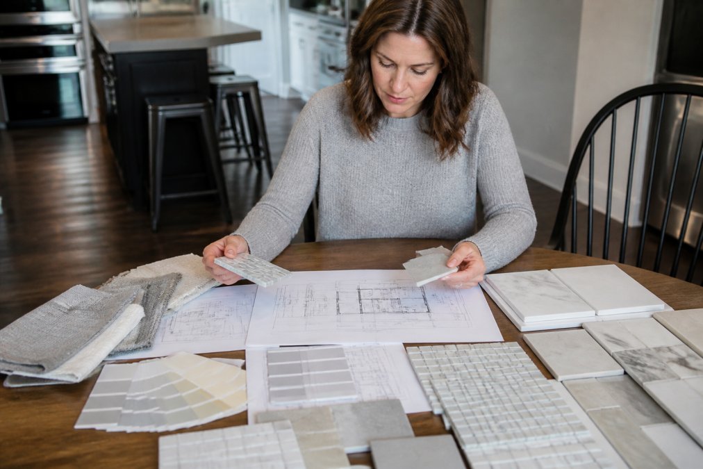 Homeowner reviewing bathroom renovation design plans and material samples at a kitchen table with tile samples, paint swatches, and floor plans spread out