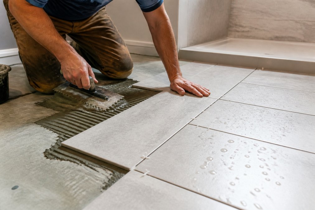 Close-up of professional installing porcelain tile on bathroom floor with proper spacing and grout application, water droplets visible on finished tiles to show moisture resistance