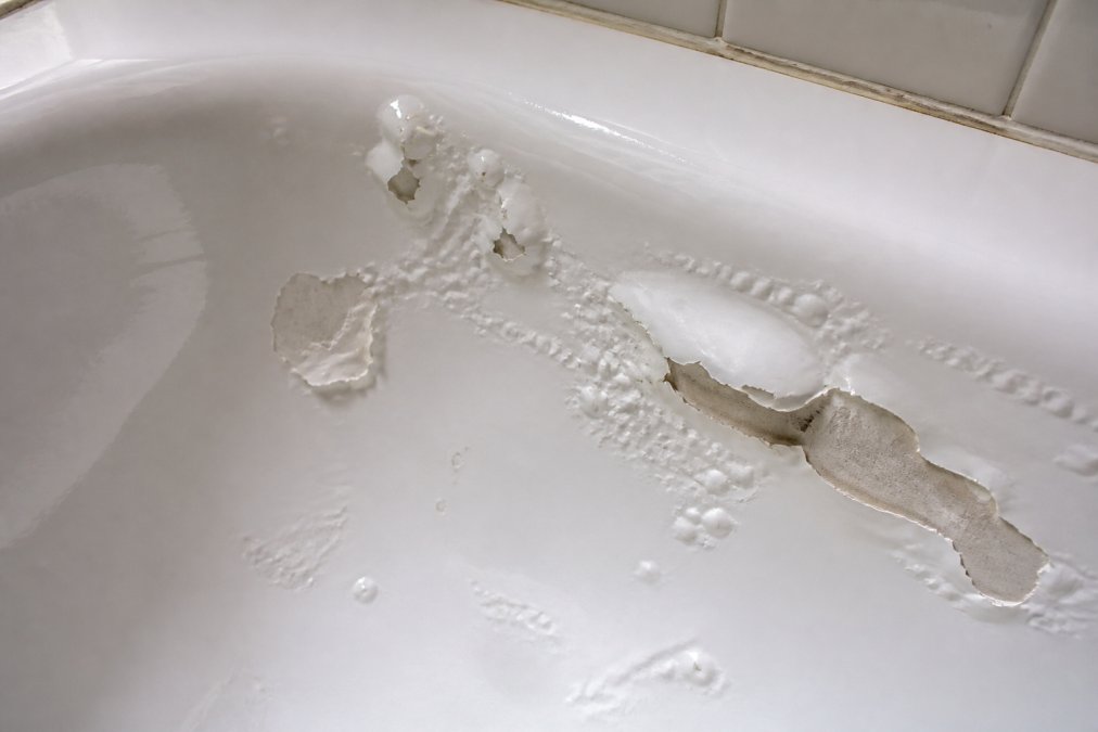 Close-up of a bathtub surface showing peeling, bubbling, and blistering on the coating with visible damage areas, natural bathroom lighting