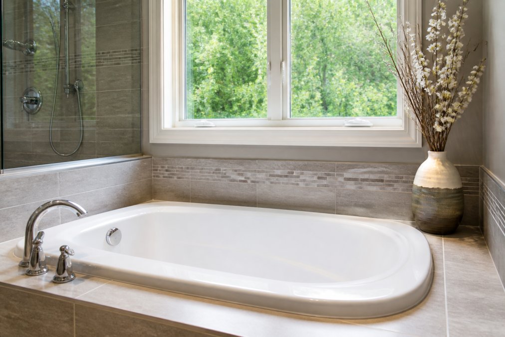 Modern bathroom featuring a gleaming acrylic bathtub with clean lines, professional tile surround in neutral tones, and polished chrome fixtures installed against a bright window