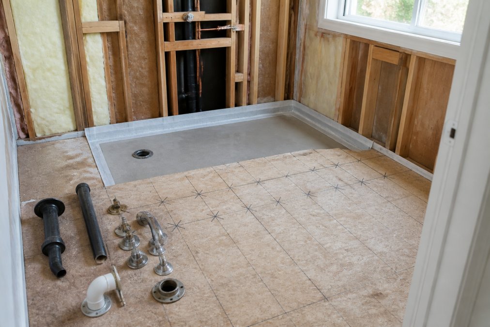 Bathroom mid-renovation showing exposed subfloor with careful inspection markings, removed plumbing fixtures laid out neatly, and fresh substrate preparation for new bathtub installation in natural daylight
