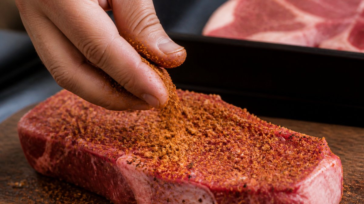 Close-up of a hand generously coating a raw steak with coarse BBQ rub, showing the texture and granules adhering to the meat surface