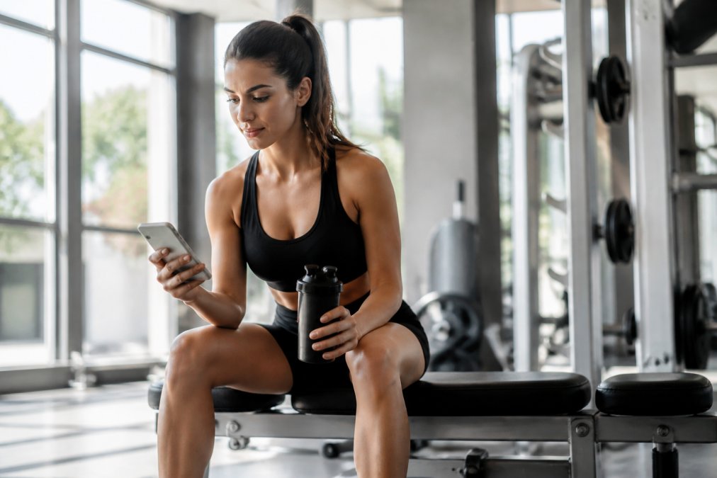A fit woman in athletic wear sitting on a gym bench, holding a shaker bottle and checking a fitness app on her phone, natural light streaming through windows
