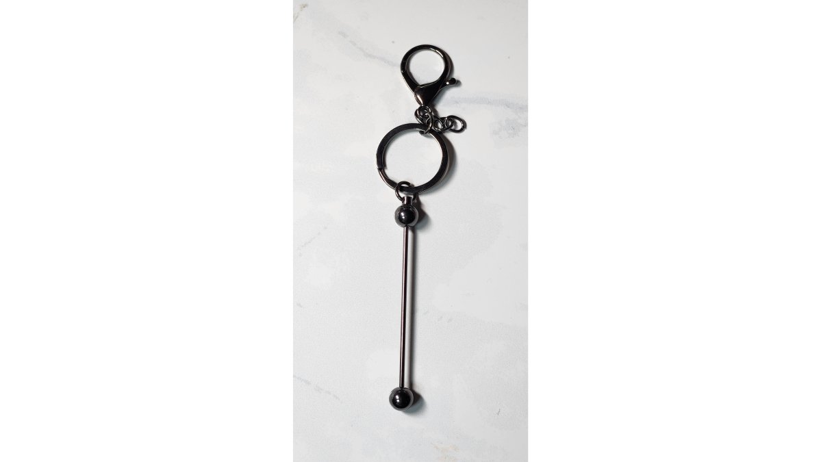 A crafter's hands threading a 15mm silicone bead onto a black nylon cord, with a beadable bar keychain and assorted beads laid out on a craft mat nearby, bright natural lighting