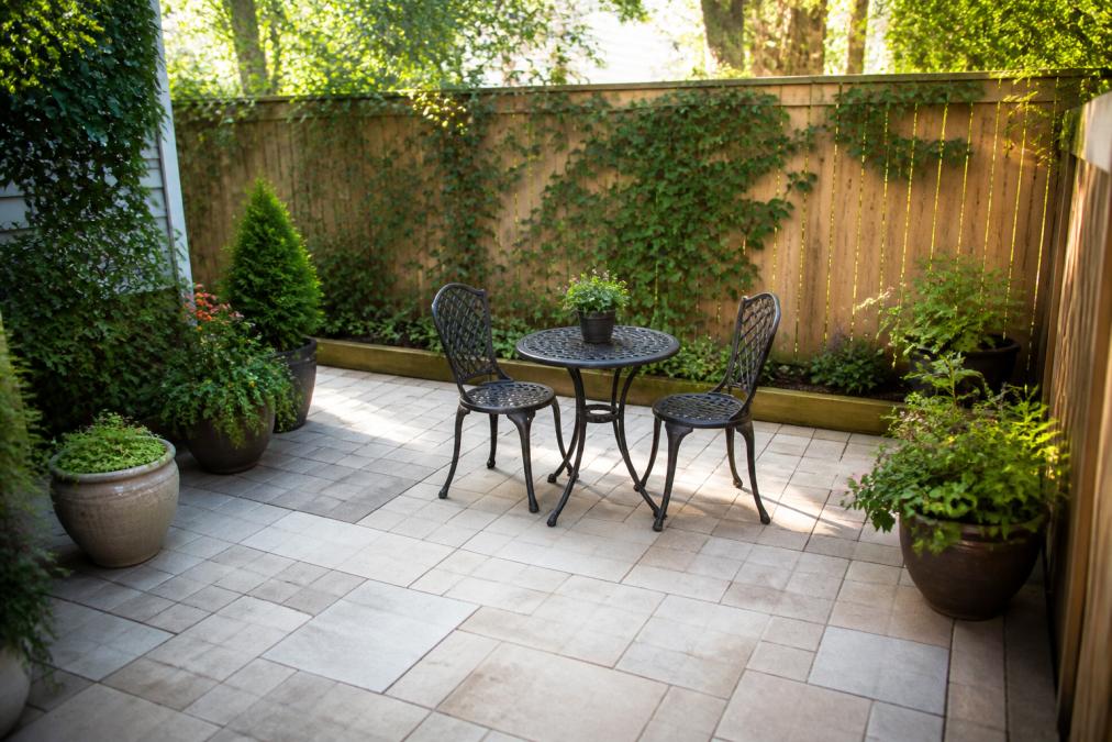 Compact residential backyard with 12-by-12-foot patio made of light-colored pavers, small bistro table with two chairs, climbing vines on wooden fence, and potted plants arranged along edges, afternoon sunlight casting soft shadows