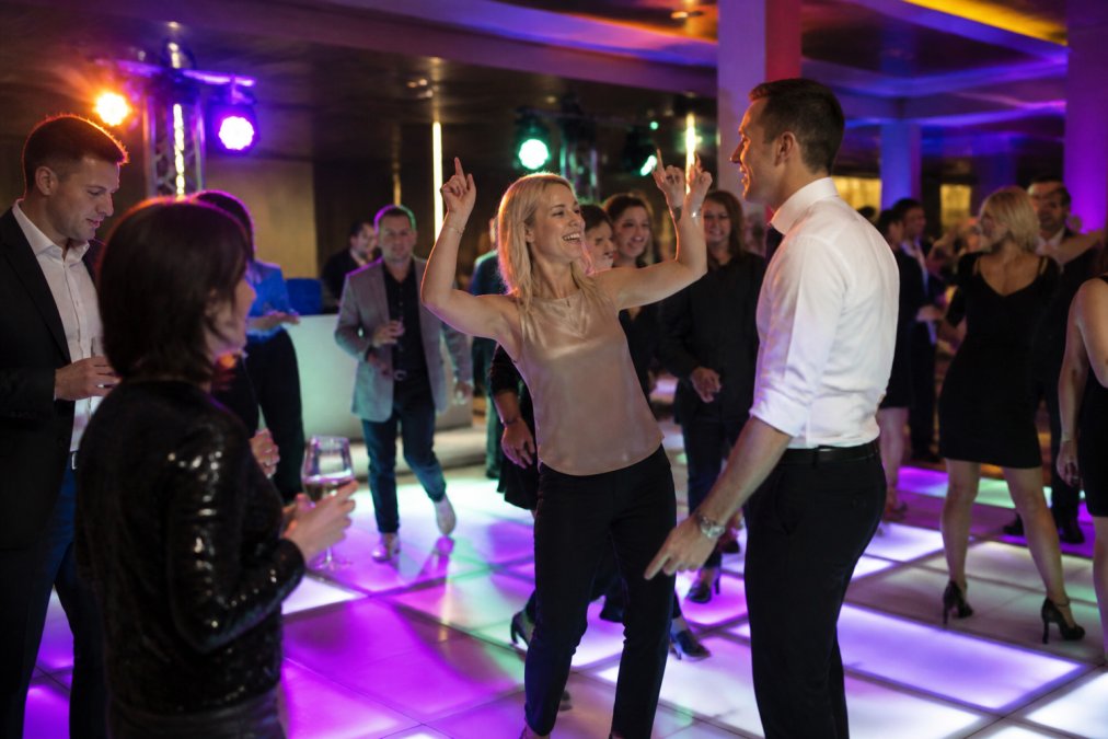 Groep collega's die enthousiast dansen op een verlichte dansvloer met kleurrijke lichteffecten tijdens een bedrijfsfeest, met vrolijke gezichten en feestelijke sfeer in een moderne zaalhuur