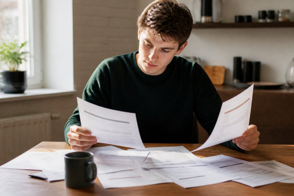Studerende sidder ved et skrivebord med laptop og papirer spredt omkring, sammenligner forskellige a-kasse-tilbud med fokus på valg og beslutningstagning i naturligt lys