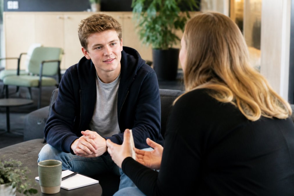 Ung person i casual tøj sidder og taler med en rådgiver i et venligt, åbent kontor-miljø, der illustrerer personlig vejledning, tillid og karrierevejledning