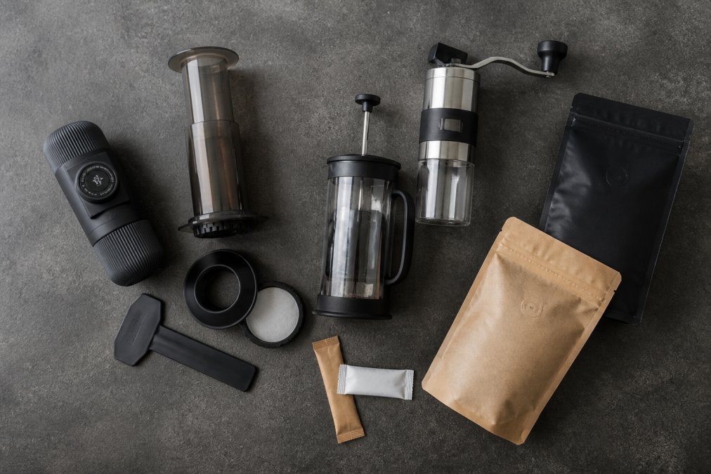 Flatlay af bærbar Nanopresso espressomaskine, AeroPress kaffebrygger, GSI JavaPress stempelkande, Petromax håndkværn, pakker med instant kaffe og specialkaffe-bønner arrangeret på en mørkegrå betonplade med naturligt dagslys