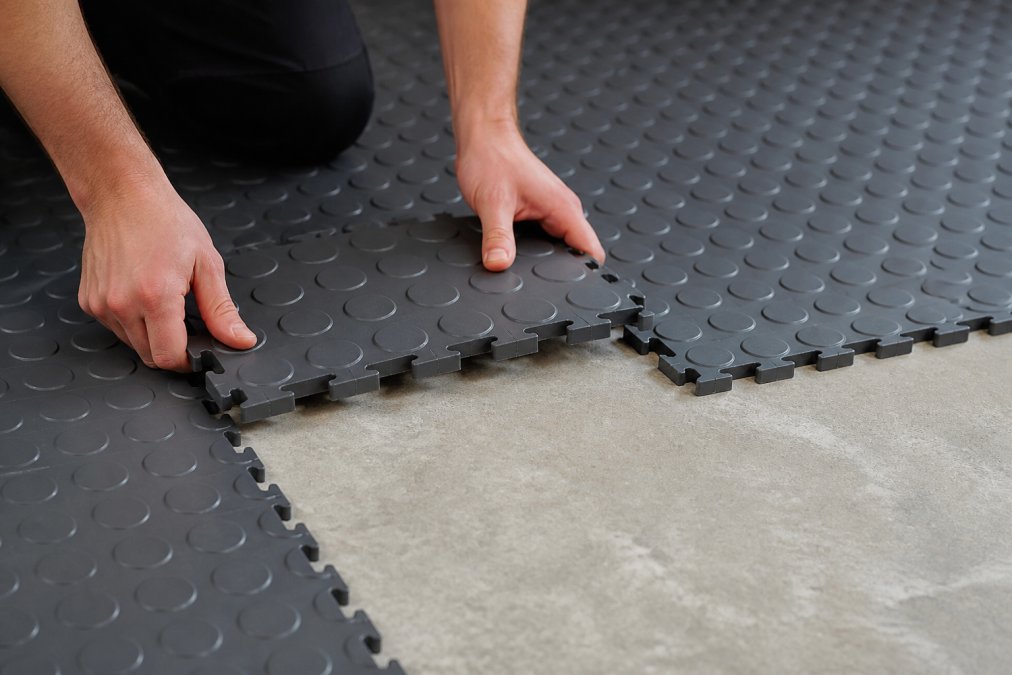 Nærbillede af interlocking PVC-gulvfliser, der bliver installeret i et garage, med en persons hænder, der positionerer fliserne på betonunderlag, som viser det modulære forbindelsessystem
