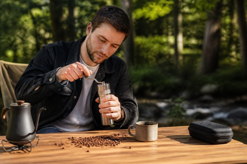 Rejsende mand sidder ved campingbord i solskin med manuel kaffekværn, kaffebønner spredt på bordet og en fyldt kaffekop, skov i baggrunden