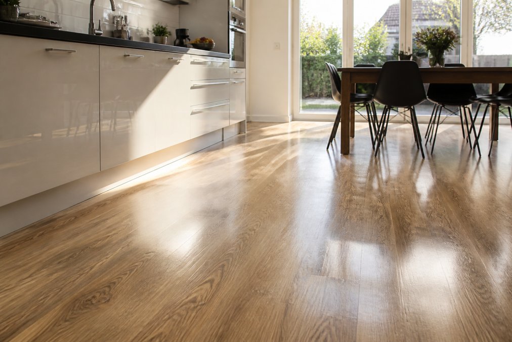 Moderne køkken med glinsende vinylgulv i træ-look, sollys reflekteres fra overfladen, spisebord og stole synlige i baggrunden