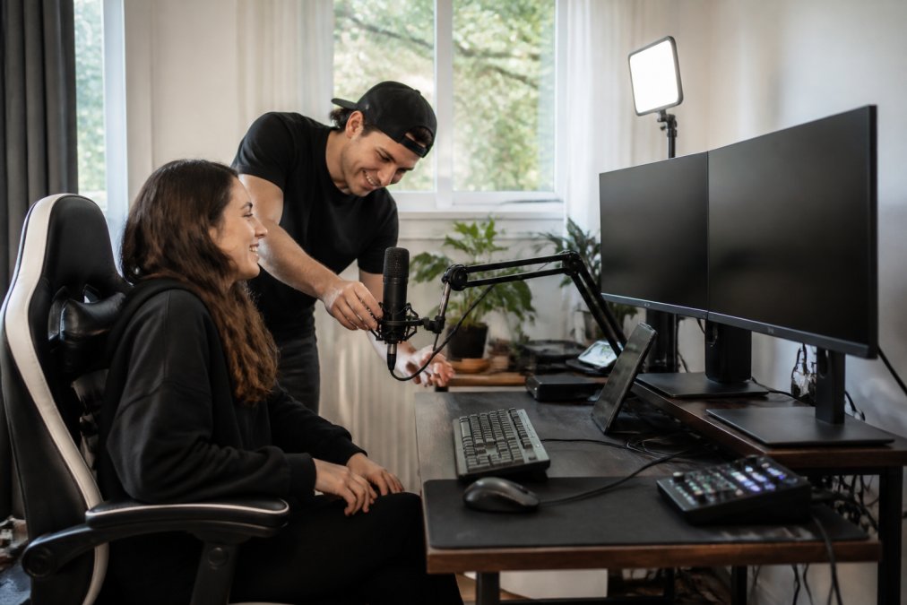 Well-organized home studio setup featuring a spacious gaming desk with dual monitor stand, ergonomic MCQ gaming chair with lumbar support, professional microphone on articulating arm, compact LED video light, and organized cable management creating a professional creator workspace with natural window lighting