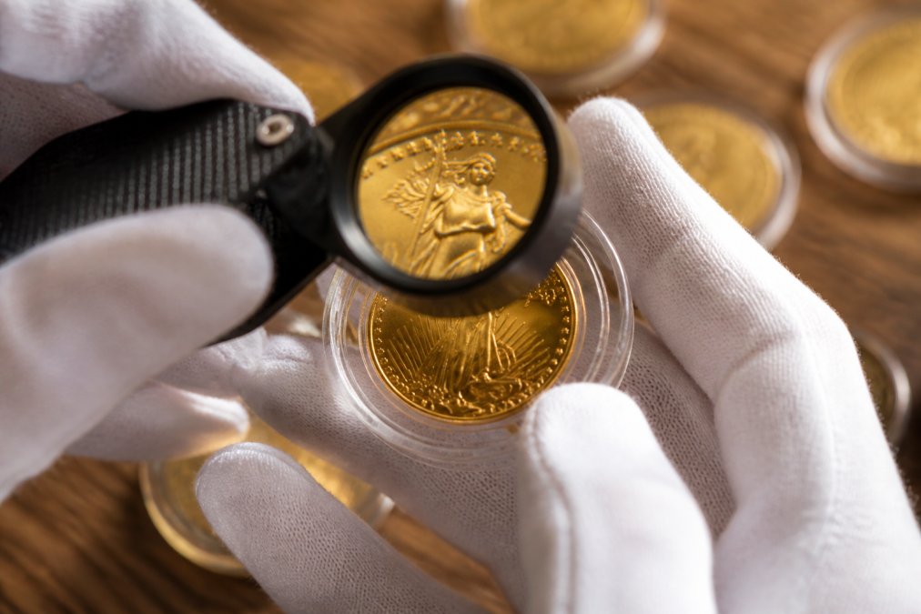 Close-up of hands holding and examining a gold coin under magnification, showing details of the coin's surface, mint marks, and luster with professional lighting