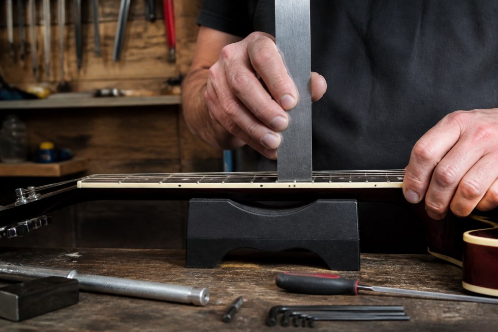 Close-up of hands holding a straightedge ruler against guitar strings at the 12th fret, measuring the gap between string and fretboard in natural workshop lighting