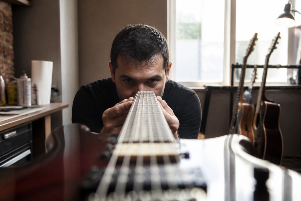 Guitarist sighting down the length of the fretboard neck toward a light source, checking for warping or uneven fret height, with guitar held at eye level in workshop setting