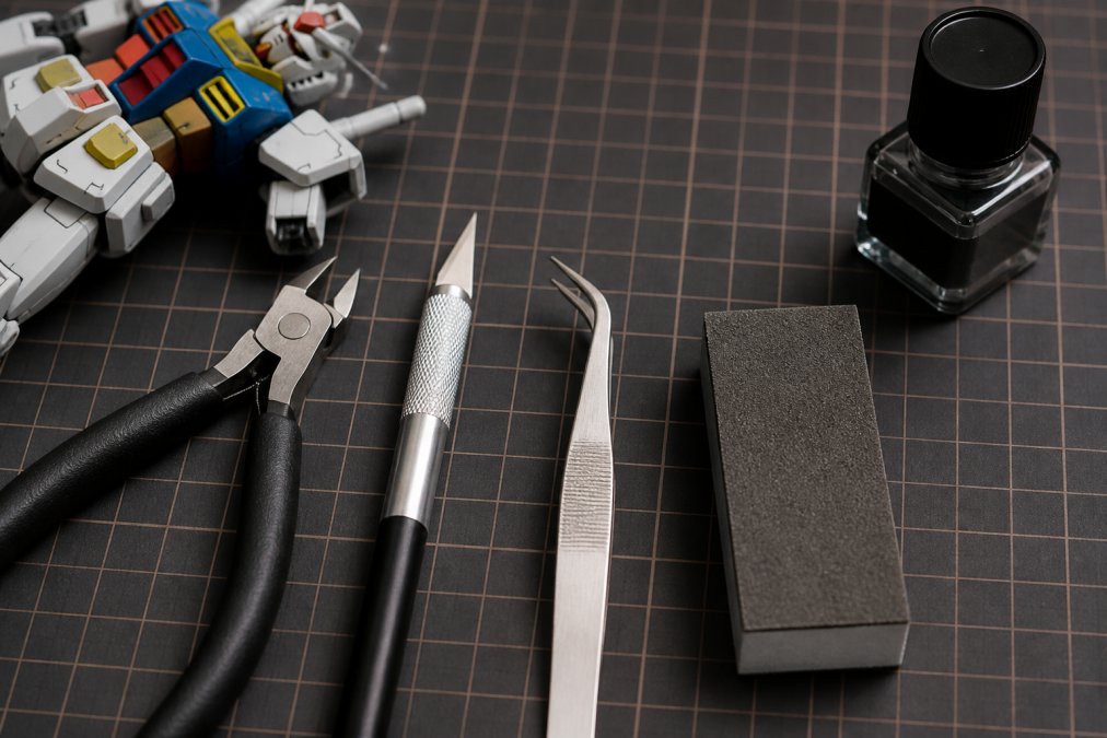 Close-up overhead shot of five essential beginner tools laid out on a work mat: side cutters, hobby knife with blade, tweezers, sanding sponge, and panel liner bottle, with a Gundam model kit part in the background