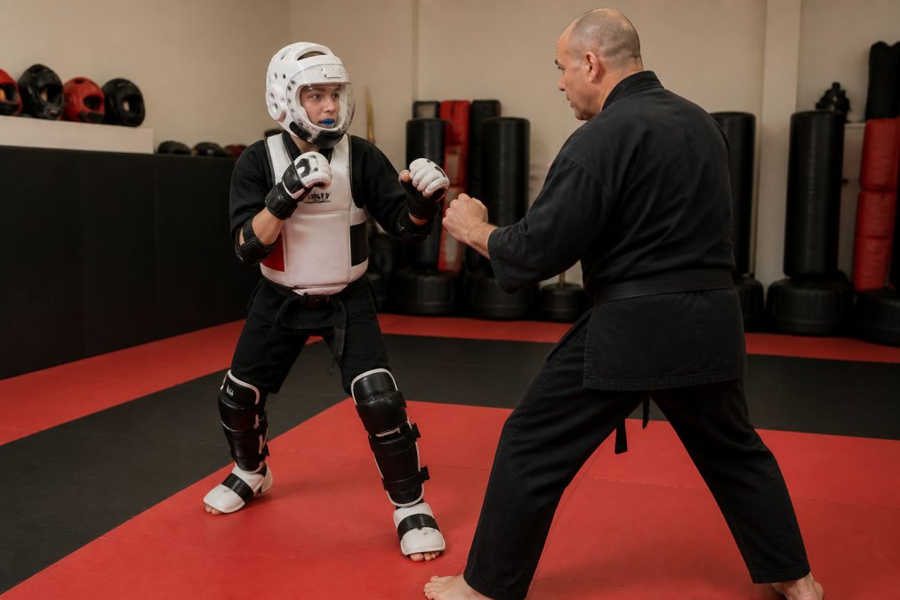 Martial artist wearing complete protective gear including white headgear with face shield, mouthguard visible, hand wraps under boxing gloves, chest protector, shin guards with foam padding, and foot protectors during controlled sparring drill with instructor