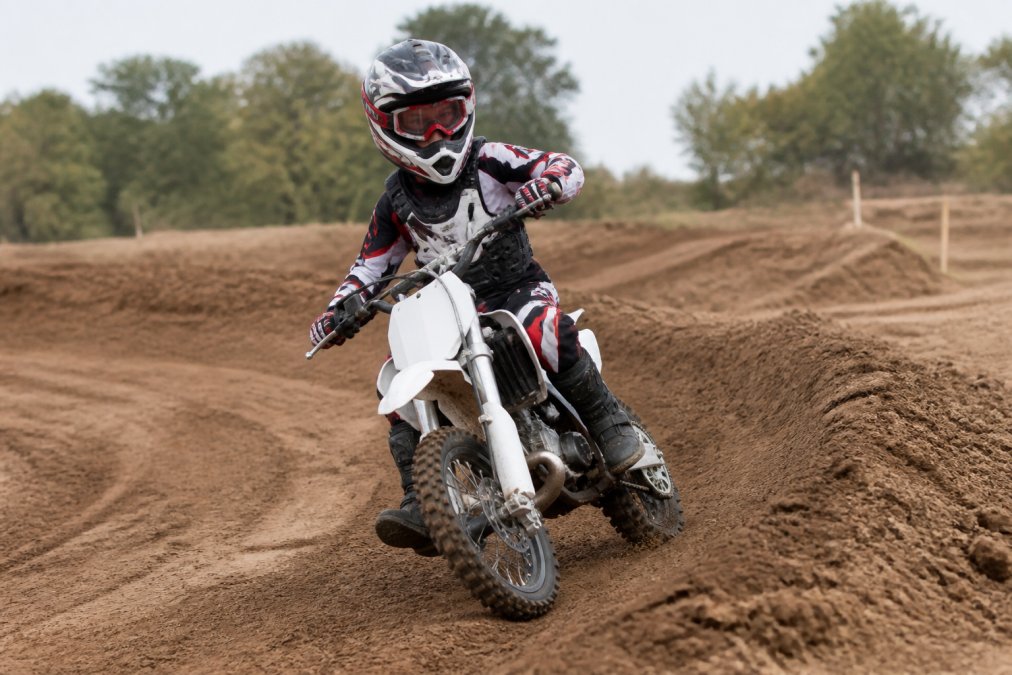 Young rider on a 50cc dirt bike demonstrating proper body positioning with bent knees gripping the fuel tank, elbows up, neutral head position looking through the turn, and hands relaxed on handlebars in a training area