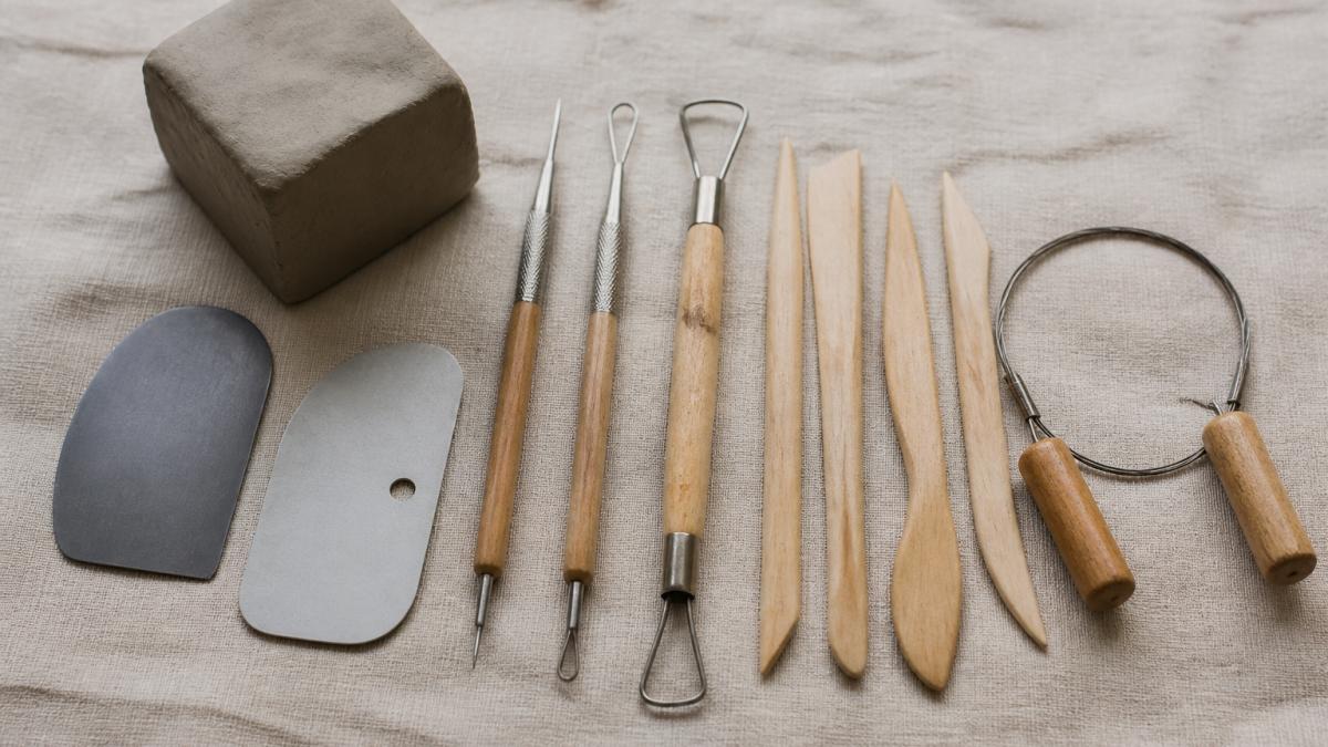 Flat-lay arrangement of beginner pottery clay tools and unfired clay on a neutral canvas surface