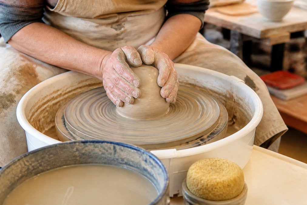 Beginner hands centering a spinning ball of clay during pottery wheel throwing in a bright studio.