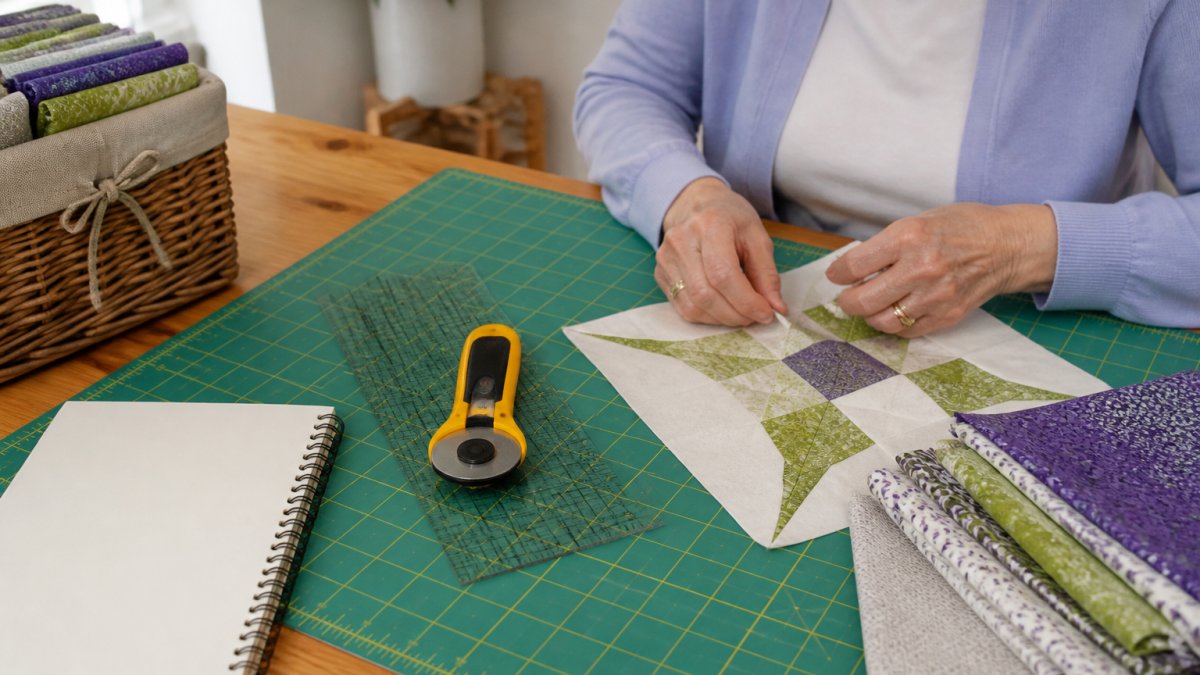 Senior woman's hands working on a quilt block with fabric, rotary cutter, and ruler on a quilting mat, with warm natural lighting