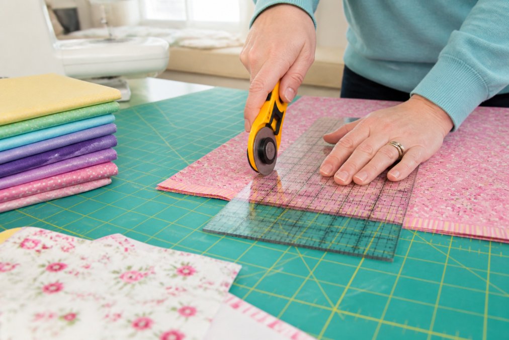 Close-up of hands using a rotary cutter and clear quilting ruler on a green self-healing mat, cutting through layers of colorful cotton fabric in a bright sewing room