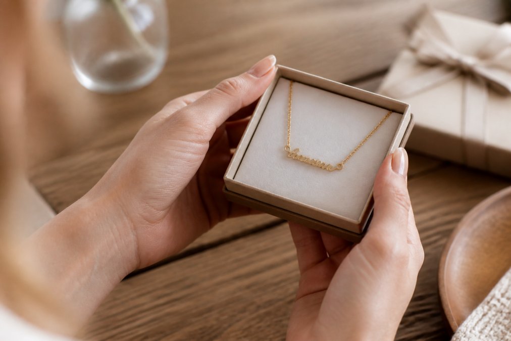 Close-up of a woman's hands holding a delicate gold name necklace in a small gift box, soft natural light on a wooden table