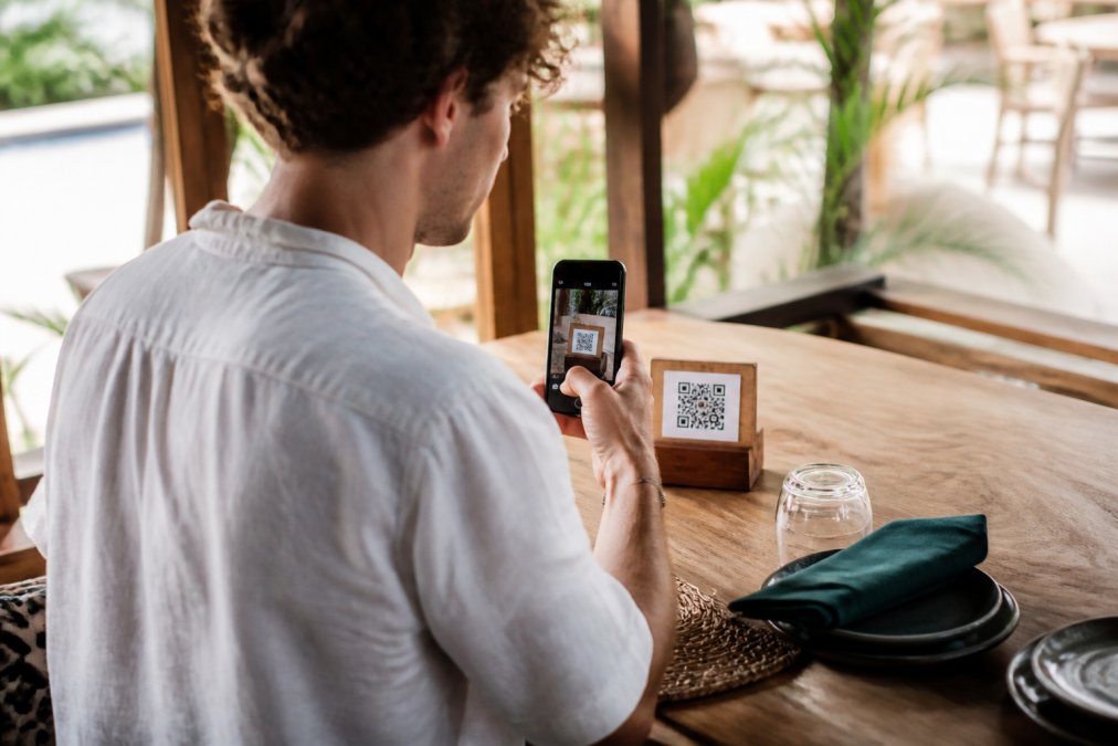 Un turista sentado en una mesa de restaurante escaneando un código QR con su smartphone para acceder al menú digital, con luz natural del restaurante iluminando la escena