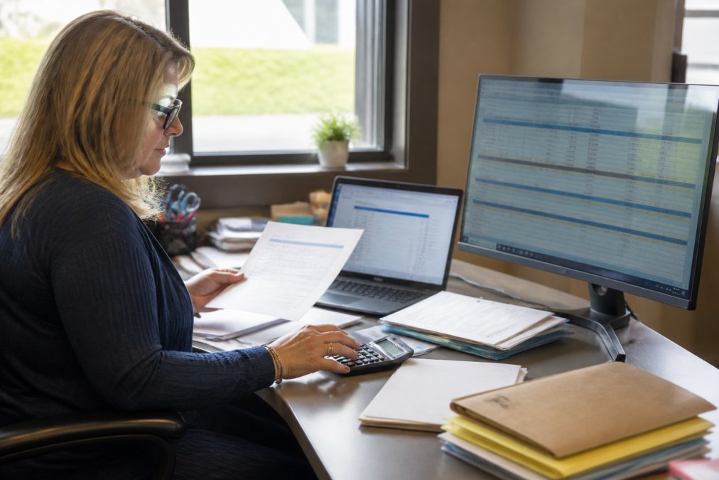 Office manager at desk reviewing organized billing documents and payroll records displayed on computer screen, with calculator, file folders, and printed statements visible in natural office lighting
