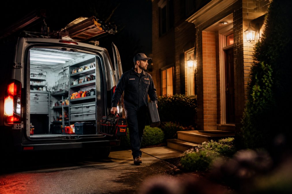 Licensed HVAC technician in uniform arriving at a residential home at night with fully stocked service truck, carrying diagnostic equipment and replacement parts, professional lighting from truck illuminating the entrance