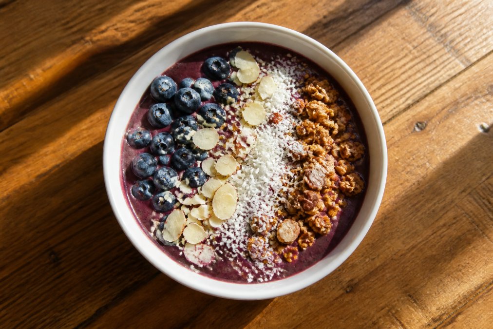 Beautifully arranged acai bowl with fresh blueberries, sliced almonds, shredded coconut, and granola scattered across a vibrant purple base, photographed from above on a wooden table with natural morning sunlight streaming across the surface