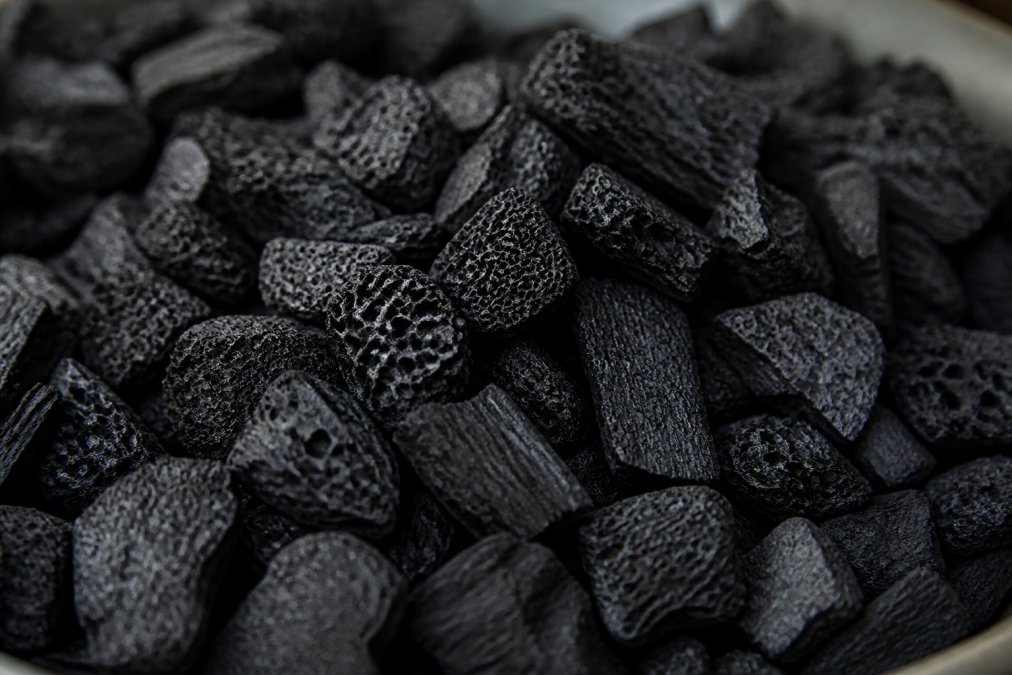 Close-up of activated carbon filter material showing dense black porous granules with intricate surface texture, natural light filtering through to highlight the extensive pore structure